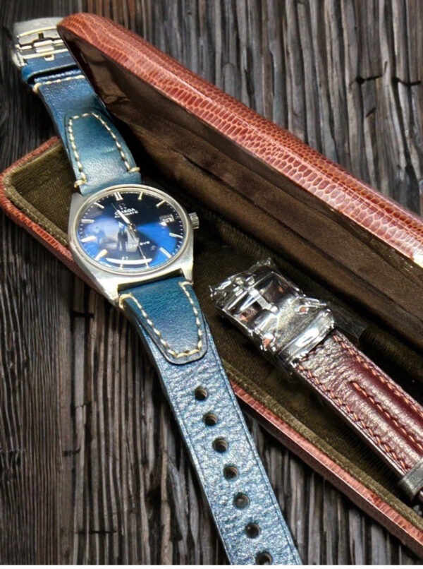 Seiko watch with a blue dial and leather strap resting on a textured wooden surface, alongside a second leather strap in a luxurious case.